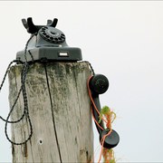 Ein altes schwarzes Wählscheibentelefon steht auf einem alten Pfeiler am Strand. Der Hörer hängt herab. In der Schnur ist Treibgut verheddert.