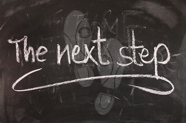 Auf einer Tafel steht mit Kreide: "The next step", im Huntergrund Fußabdrücke aus Kreidestaub.