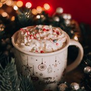 Roter Hintergrund mit unscharfen Lichtern. Im Vordergrund Tannenzwige um eine Tasse Kakao mit Sahne und Zuckerperlen.