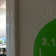 Weiße Wand und Tür mit einem Auschnitt eines grünen Schildes auf dem 2.10 steht. Durch die Tür, leicht verschommen, ein Bürostuhl und Schreibtisch