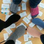 Auf einem Holzdielenboden liegen viele EUTB Visitenkarten durcheinander. Darauf stehen drei Personen. Man sieht ihre Füße in Socken. Jede Soche hat eine andere Farbe.