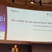 Mathias Teller am Rednerpult auf der Bühne. Auf einer großen Leinwand steht "DAs Leitbild als das gemeinsame Beste - Themeneinführung".