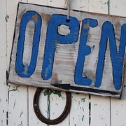 Ein weißes Holzschilz vor einer weißen Holzwand mit blauer Schrift: OPEN