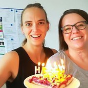 Frau Starke und Frau Siemens-Gerth halten gemeinsam den Kuchen mit brennenden Kerzen in die Kamera. Sie lachen.
