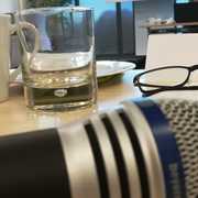 Nah fotografiert: Ein Mikrophon auf einem Tisch. Im Hintergrund ein Wasserglas und eine Brille. Restlicher Hintergrund verschwommen. 