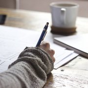 Bildausschnitt. Schreibtisch. Die recht Hand einer Frau hält einen Stift. Auf dem Tisch Unterlagen. Im Hintergrund verschwommen eine Tasse.