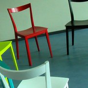 Bildauschnitt. Sechs Stühle im Kreis. Zwei Davon angeschnitten. Die Stühle sind aus Holz und bunt lackiert in weiß, türkis, gelb, rot schwarz und gelb.