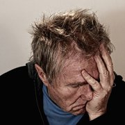 Vor neutralem Hintergrund. Portrait eines älteren Mannes mit zerzaustem Haar. Er vergräbt das Gesicht verzweifelt in seiner linken Hand.